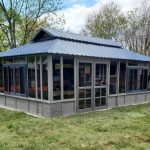 A large gray gazebo with a metal roof set on a grassy lawn, surrounded by trees and fencing.