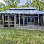 A gray metal-roofed gazebo with glass windows sits on a grassy lawn with trees in the background.