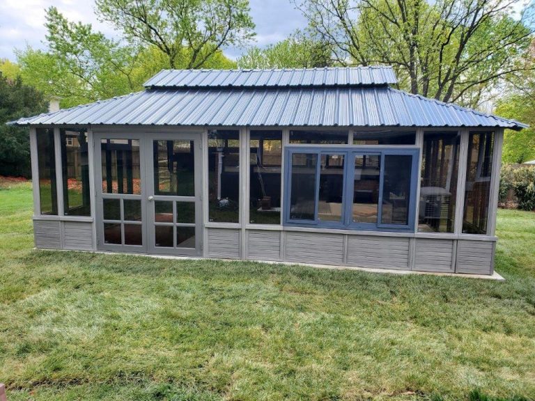 A gray metal-roofed gazebo with glass windows sits on a grassy lawn with trees in the background.