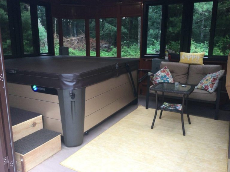 Indoor hot tub with steps, next to a sofa with colorful pillows, a small table, and a rug in a room with large windows.
