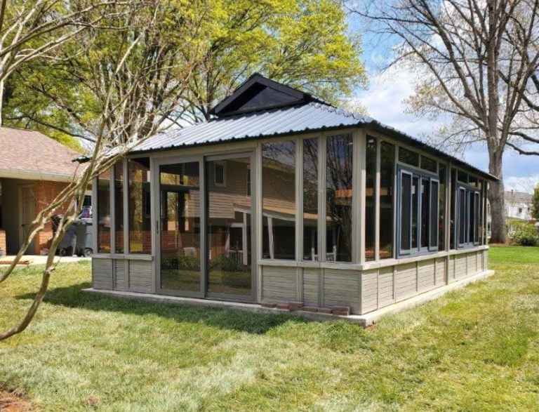 A sunroom with large windows and a metal roof in a backyard surrounded by trees and grass.