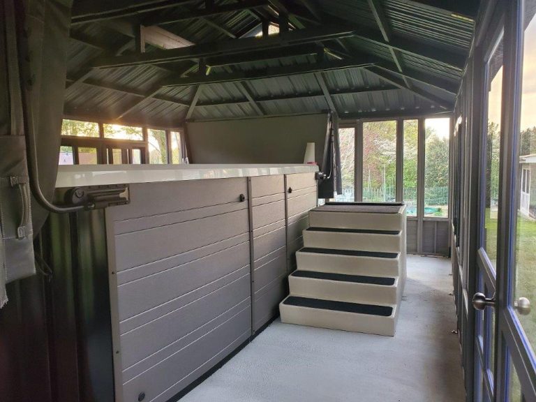 Indoor gazebo with glass walls, a covered hot tub, and steps under a metal roof, overlooking a garden.