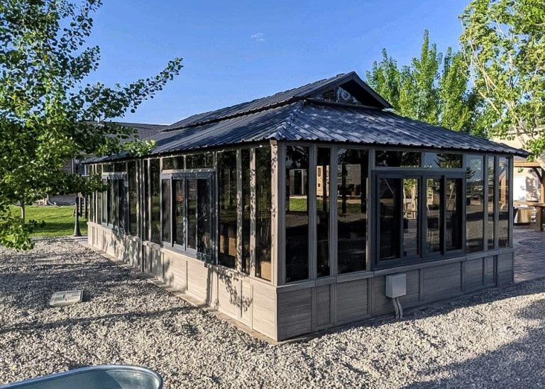 A glass-walled pavilion with a dark roof is surrounded by gravel and trees under a clear blue sky.