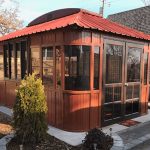 Red-roofed wooden gazebo with glass walls, set on concrete, surrounded by small plants and trees.