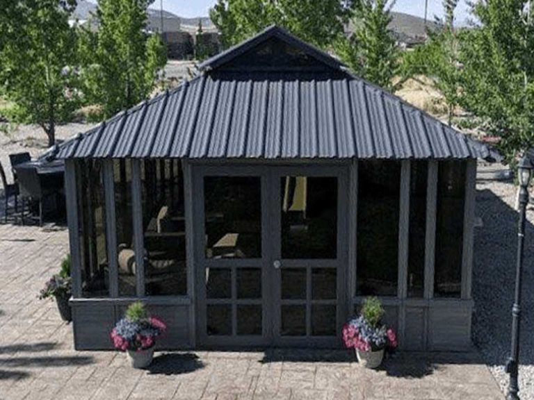 Gray garden gazebo with glass panes, surrounded by potted flowers and trees on a stone patio.