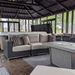 Modern sunroom with gray wicker sofas, glass fire table, hot tub, and large windows showing trees outside.