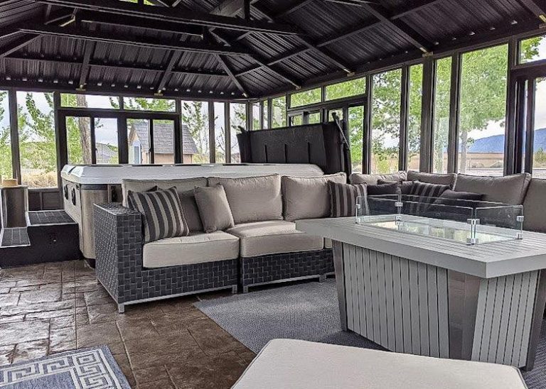 Modern sunroom with gray wicker sofas, glass fire table, hot tub, and large windows showing trees outside.
